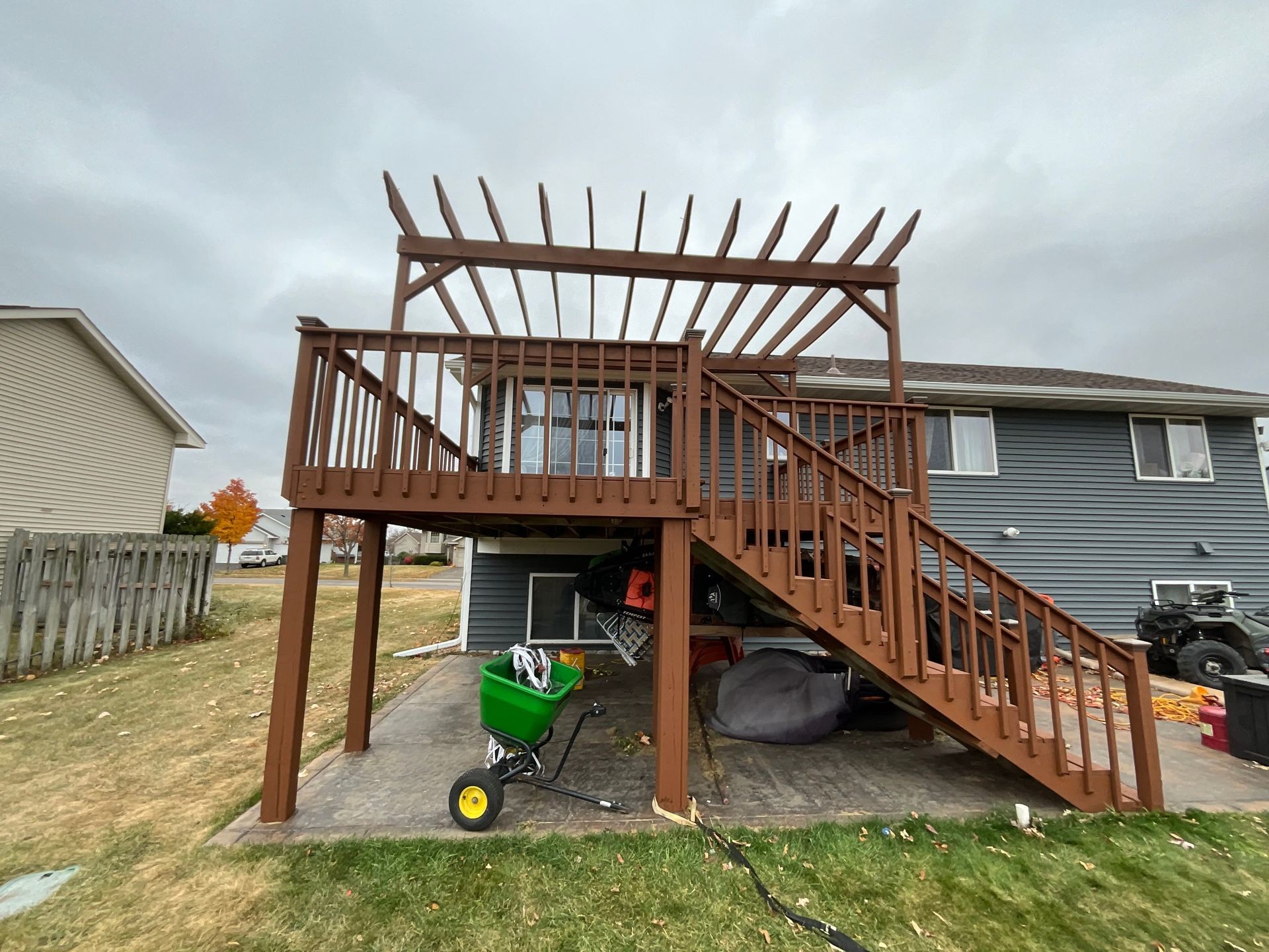 Two-level wooden deck with stairs and pergola attached to a house, with lawn equipment beneath.
